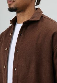 Bruine overhemdjas van een wolmix met een kraag, voorzien van knoopsluitingen en een zachte textuur, gedragen over een wit T-shirt.