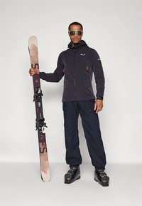 Suusapüksid, millel on must lukuga jope oranžide detailidega, mereväe sinised püksid ja mustad suusasaapad. Hoidmas pruune suuski koos kinnitustega.