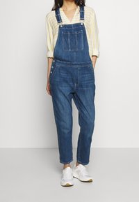 Mörkblå denimbyxor med justerbara remmar, framficka, sidoknappar och tapered bendesign. Matchas med vita sneakers.