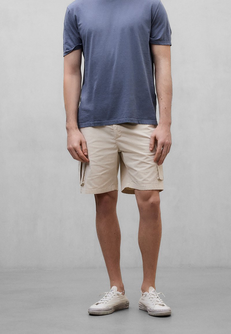 Homme portant un t-shirt bleu, un short cargo beige et des baskets blanches, debout devant un fond gris clair uni.