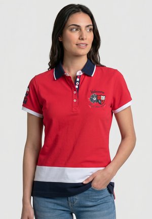 Mujer con polo rojo con cuello azul marino, ribete blanco y azul marino en el bajo y escudo bordado, de pie con la mano en el bolsillo del vaquero.