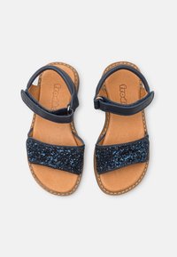 Marineblå sandaler med en glitrende blå rem, læderindlæg og justerbar ankelrem. Glat tekstur med syede kanter.