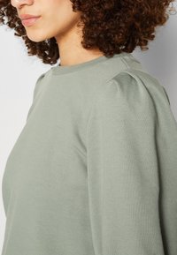 Lys grøn pullover med en glat tekstur, der har samlet detaljer på skuldrene og en rund halsudskæring.