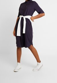 Person klädd i en mörkblå knappklänning med en vit tröja knuten runt midjan och vita högklackade sneakers som står mot en vit bakgrund.