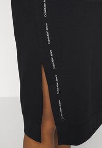 Mustapohjainen polvipituinen hame, jossa on sivureikä, ja jonka reunalla kulkee pystysuora valkoinen "Calvin Klein Jeans" -teksti.