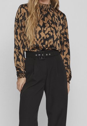 Vrouw draagt een bruine en zwarte blouse met dierenprint en lange mouwen, gestoken in een zwarte broek met hoge taille en een brede riem.