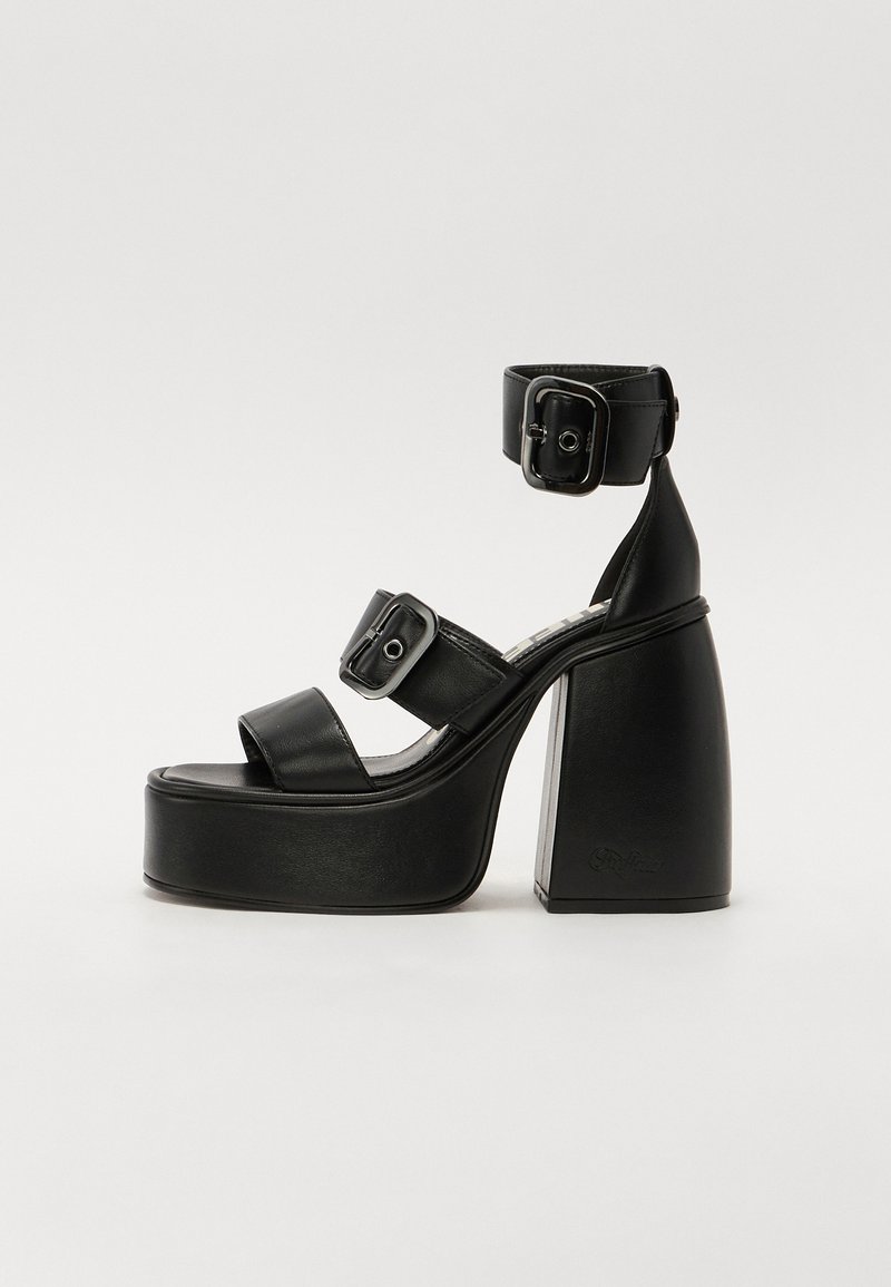 Sandálias de plataforma pretas com um salto robusto, apresentando três tiras largas e uma tira no tornozelo com fivela. Feitas de um material liso semelhante ao couro.