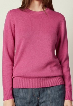 Vrouw die een effen felroze trui draagt met lange mouwen en geribde boorden, gecombineerd met een blauwe spijkerbroek.