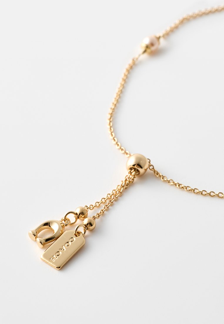 Collier en or comprenant une fine chaîne, des perles rondes et deux pendentifs suspendus : une boucle et une étiquette rectangulaire avec un texte gravé.