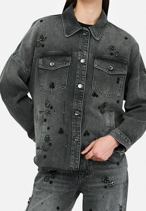 Sort denimjakke og jeans med sorte perlebesatte blomsterudsmykninger båret af person med hånden i lommen.