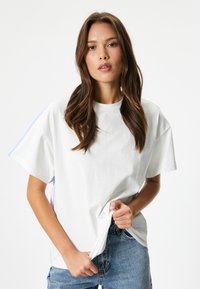 Witte oversized T-shirt met korte mouwen en een ronde halslijn, met lichtblauwe accenten op de schouders en zijkanten. Draagt met lichte spijkerbroek.