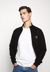 Svart zip-up jacka med hög krage, texturerad tyg och ett litet logopatch. Bärs över en vit t-shirt, tillsammans med blåjeans.