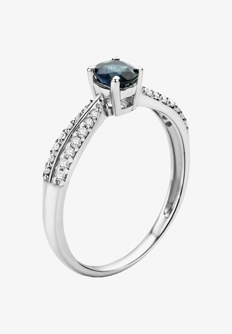 Anillo de compromiso de plata con una gema azul ovalada rodeada de pequeñas piedras transparentes a lo largo de la banda, con un acabado elegante y pulido.