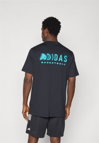 Zwarte T-shirt met korte mouwen, voorzien van een turkooise stippenpatroon met het "ADIDAS BASKETBALL" logo op de achterkant; gecombineerd met zwarte shorts.