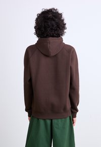 Brown hoodie with a large hood, raglan sleeves, and ribbed cuffs. Paired with green pants, showing soft fabric texture on both garments.
