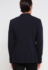 Man som bär en mörk marinblå kavaj med tunna horisontella ränder, sedd bakifrån mot en enkel ljus bakgrund.