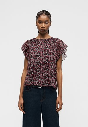 Jeune femme aux cheveux tressés portant un chemisier à manches courtes à motif floral avec des manches à volants et un pantalon large sombre, devant un fond uni.