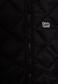 Sort quiltet jakke med en central lynlås og et lille hvidt broderet "Lee" logo på højre side.