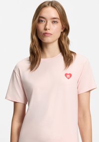 Ροζ ανοιχτόχρωμο t-shirt με κοντά μανίκια, κατασκευασμένο από απαλό ύφασμα, με κόκκινο λογότυπο καρδιάς και το κείμενο "HUGO" στην αριστερή πλευρά του στήθους.