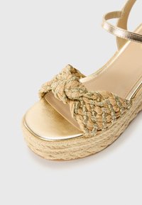 Hælestropp sandal med en gullmetallisk stropp og flettet naturfiber på toppen. Har en tykk vevd jute såle og en glatt, skinnende tådel.