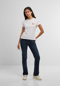 Witte katoenen t-shirt met een rood logo op de borst, gecombineerd met donkerblauwe spijkerbroek en witte sneakers, staande tegen een neutrale achtergrond.