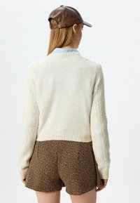 Crèmekleurige kabelgebreide trui met ribbels aan de onderkant, gecombineerd met broekshorts in bruin houndstooth. Het model draagt een bruin leren pet en een paar oorringen.