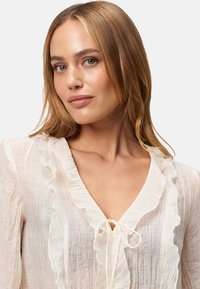 White, lightweight blouse with ruffled neckline and tie detail. Textured fabric with sheer appearance, featuring a subtle crinkle design.