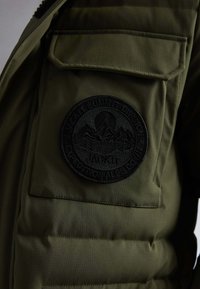 Oliwkowa kurtka puchowa z dużym czarnym okrągłym logo z górami. Tkanina ma subtelną teksturę i widoczne detale szycia.