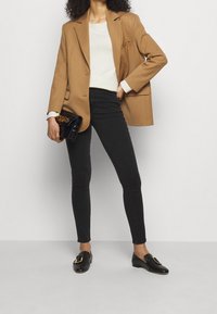 Blazer marrón sobre una blusa blanca, jeans skinny negros y mocasines negros con una hebilla dorada. Sosteniendo un pequeño bolso negro con un acabado texturizado.