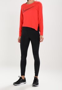 Czerwony skrócony sweatshirt z czarnym logo Nike, w połączeniu z czarnymi legginsami z teksturowanymi panelami, uzupełniony czarnymi butami sportowymi.