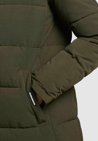 Olivengrøn pufferjakke med quiltet tekstur, der har en sidelomme og manchetterede ærmer. Stoffet er glat, let og isoleret.