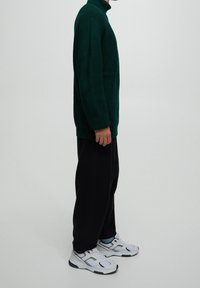Personne portant un pull en tricot vert foncé, un pantalon noir ample, des chaussettes bleu clair et des baskets blanches, debout de profil sur un fond uni.