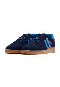 Navy blue suede sneakers with blue accents, white stripes, and a light brown rubber sole. Features flat laces and a padded collar.