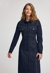 Mörkblå denimskjortklänning med långa ärmar, två bröstfickor med knappsmyckning och kontrasterande sömnadsdetaljer längs sömmarna.
