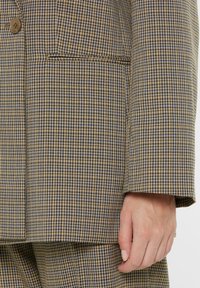 Checkered blazer featuring a brown and blue pattern, single button closure, notch lapel, and a pocket on the left side.