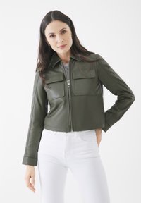 Olivgrüne Lederjacke im Cropped-Stil mit zwei Brusttaschen, Reißverschluss und Kragen, kombiniert mit weißen Jeans.