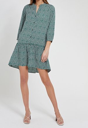 Kvinde iført en grøn kjole med stjernemønster, knaplukning, lange ærmer, udvidet kant og hvide, åbne hælede sandals.