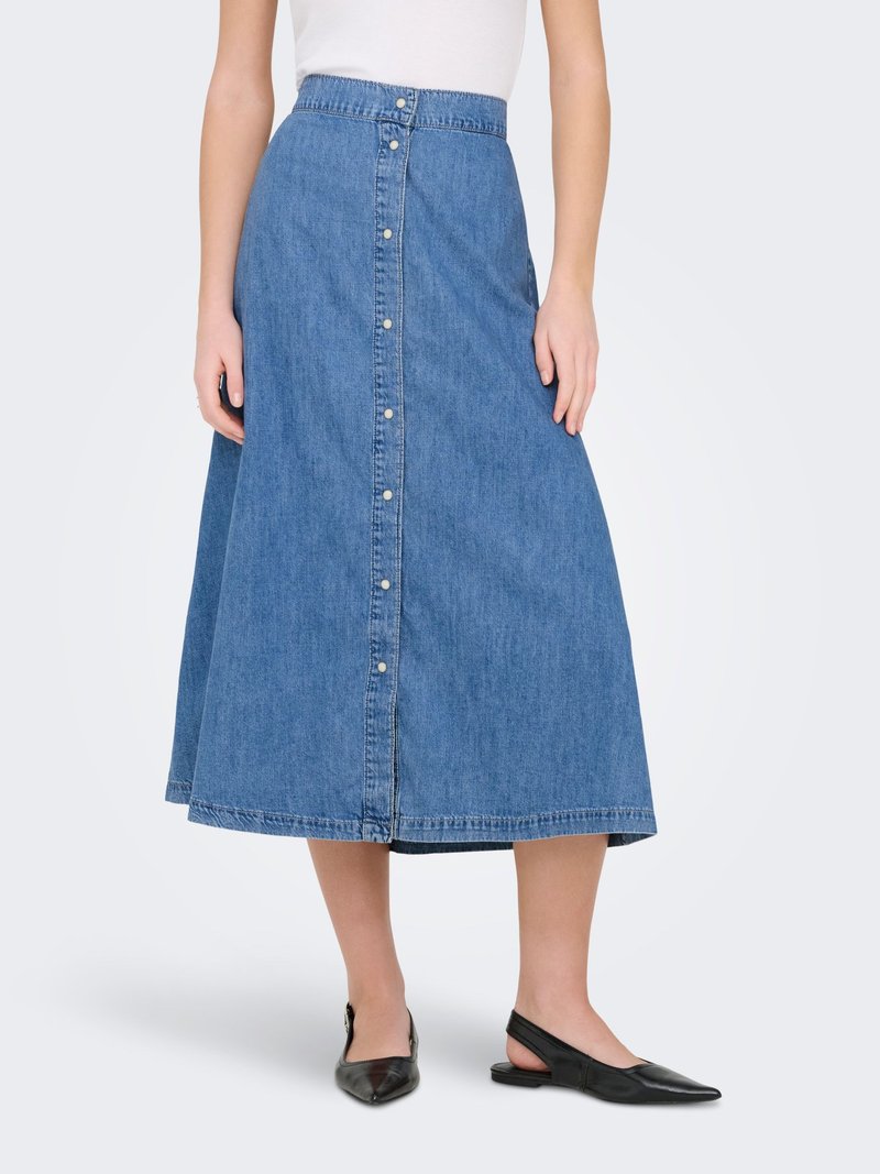 Femme portant une jupe midi en jean bleue à boutons sur le devant, avec des ballerines noir à bout pointu et à bride arrière, debout devant un fond uni.
