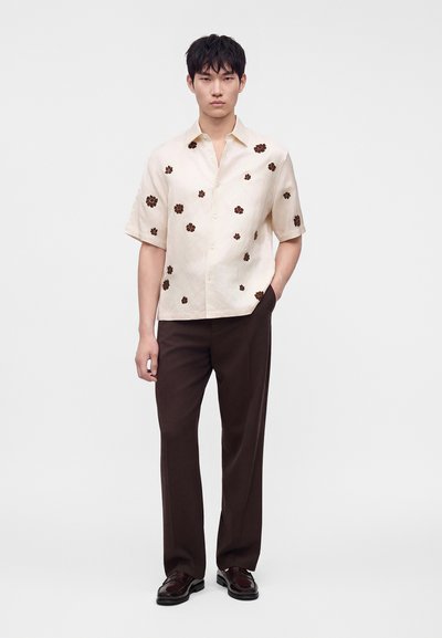Homme portant une chemise beige à manches courtes avec des motifs floraux marron, un pantalon marron foncé et des chaussures habillées marron foncé, debout avec une main dans la poche.