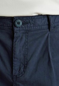 Ceinture de pantalon bleu marine avec un bouton à quatre trous étiqueté "Joules" et une passants de ceinture visible, porté avec un haut de couleur claire.