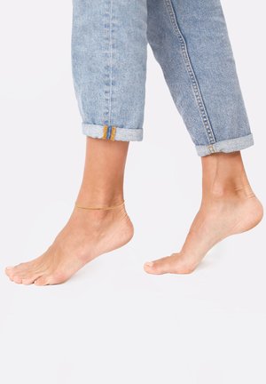 Kuldne kett anklet, mis on kantud paljastatud pahkluul, koos kergete siniste teksadega, millele on pandud üles. Sile ja läikiv tekstuur.