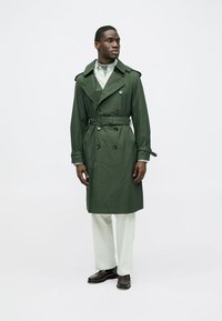 Green waterproof trench coat with double-breasted buttons, wide collar, and tied belt. Paired with light-colored trousers and brown shoes.