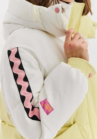 Padded bunda v bielej a pastelovej žltej farbe, s ružovým vlnkovým dizajnom na rukáve a textúrovanou látkou s ružovým logom.
