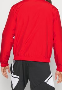 Rød vindjakke med elastiske manchetter og kant, der har en glat, blank tekstur; båret over sorte shorts med hvide accenter.