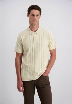 Mann trägt cremefarbenes kurzärmeliges Poloshirt und dunkelbraune Hose, steht mit einer Hand in der Tasche vor hellem einfarbigem Hintergrund.