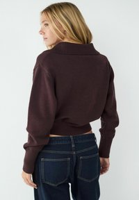 Burgunderfarbener, gekürzter Pullover mit einem breiten gerippten Kragen und langen Ärmeln, kombiniert mit dunkelblauen, hochgeschnittenen Jeans mit Gesäßtaschen.