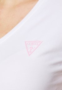 White v-neck t-shirt with a ribbed texture, featuring a small pink triangular "GUESS?" logo on the left side near the neckline.