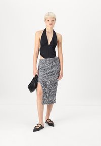 Crni halter top s dubokim V-izrezom, uparen s olovkom suknjom od srebrnih šljokica koja ima bočni prorez. Manekenka drži crnu torbu i nosi crne balerinke.