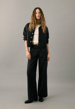 Chaqueta de mezclilla negra con botones al frente y pantalones anchos de mezclilla negra. Llevados sobre una camiseta blanca de canalé, creando un look en capas.