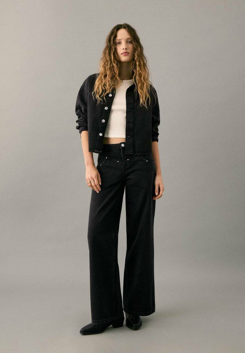 Čierna rifľová bunda so zapínaním na gombíky vpredu a čierne rifľové nohavice so širokými nohavicami. Nosí sa cez biely rebrovaný top, čo vytvára vrstvený vzhľad.